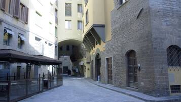 A narrow street with buildings on either side, featuring balconies, a covered seating area, and stone facades.