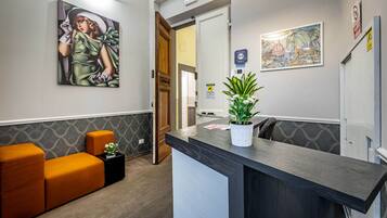 A reception area with a reception desk, a potted plant, a seating area with orange cushions, and two artworks on the walls.