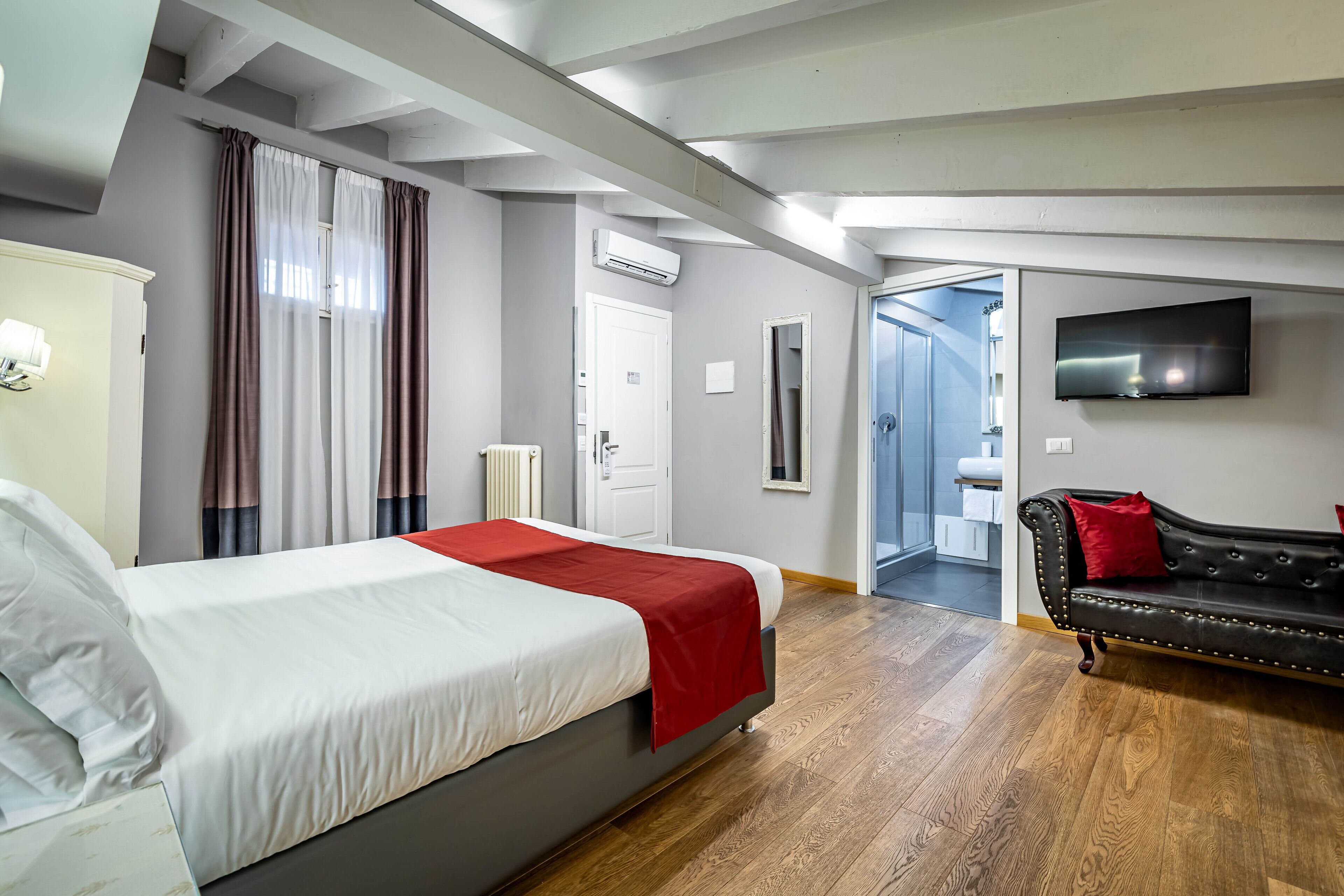 A hotel room with a bed, a red throw, a black leather sofa with a red pillow, a TV, and a bathroom visible through an open door.