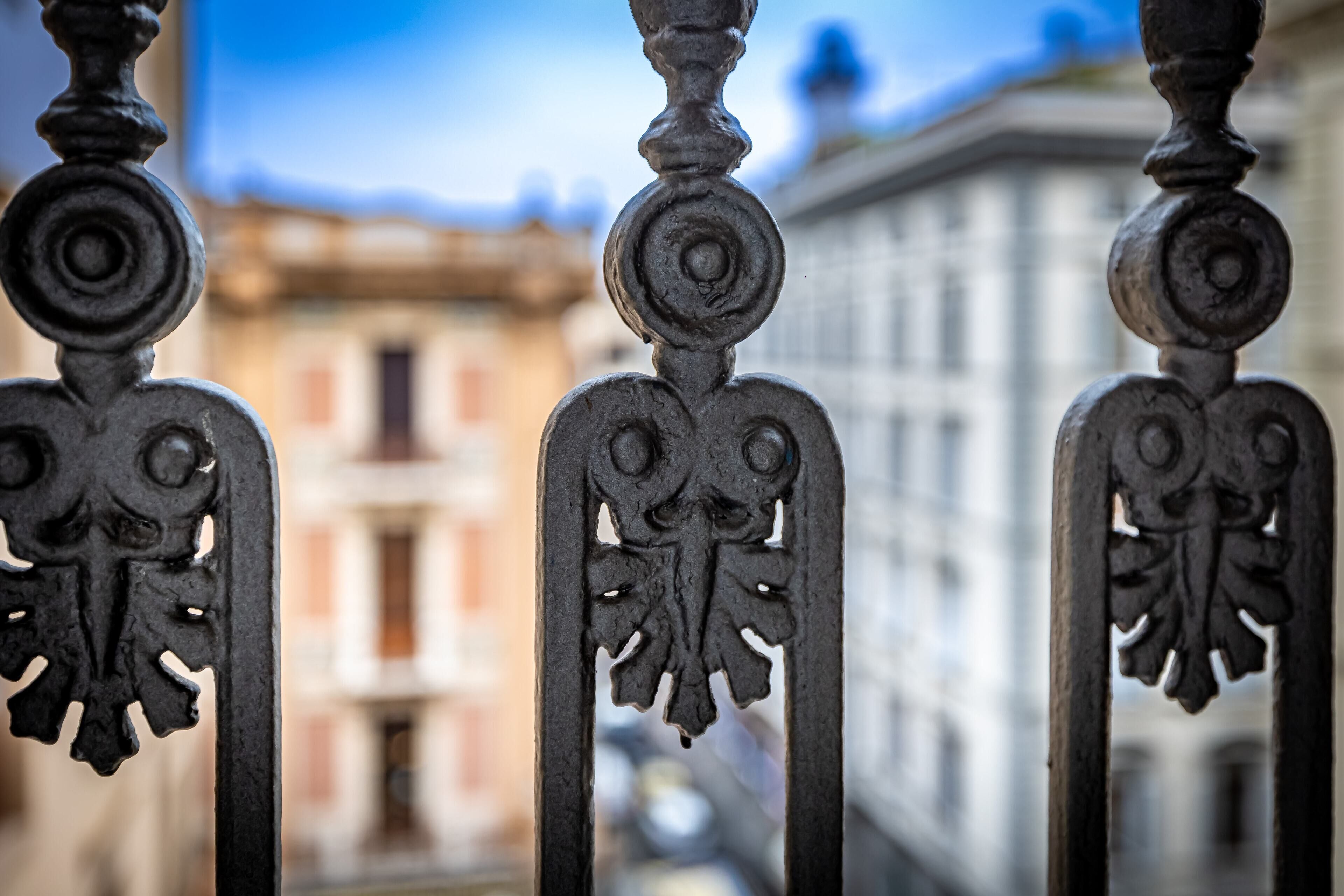A wrought iron balcony railing with intricate designs, overlooking a building with classical architecture.