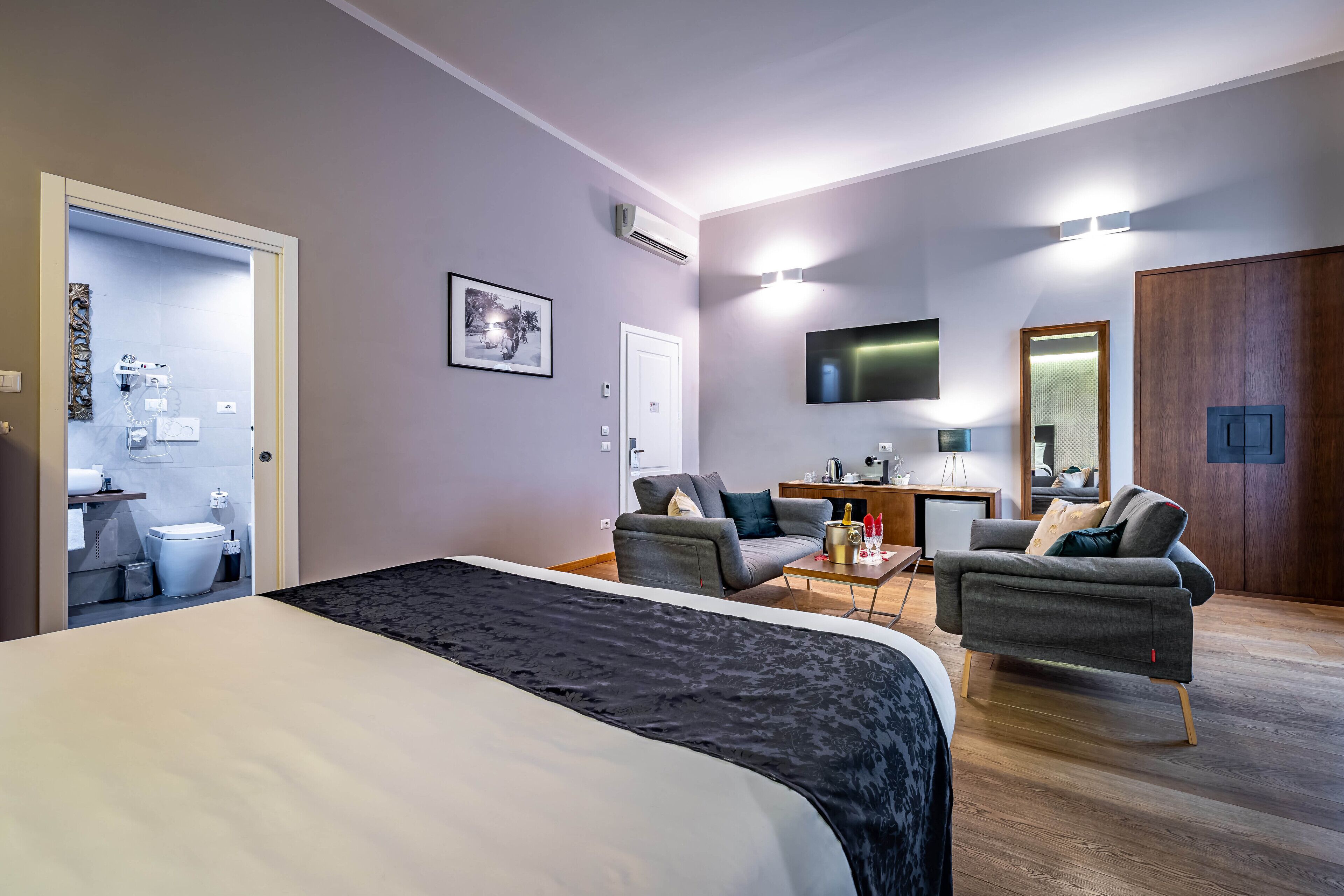 A modern hotel room with a large bed, two armchairs, a small desk, and a television.