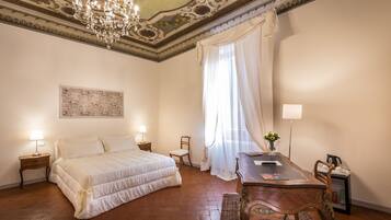 A bedroom with a large bed, a desk, and a chair. There is a chandelier, a window with curtains, and a painting on the wall.