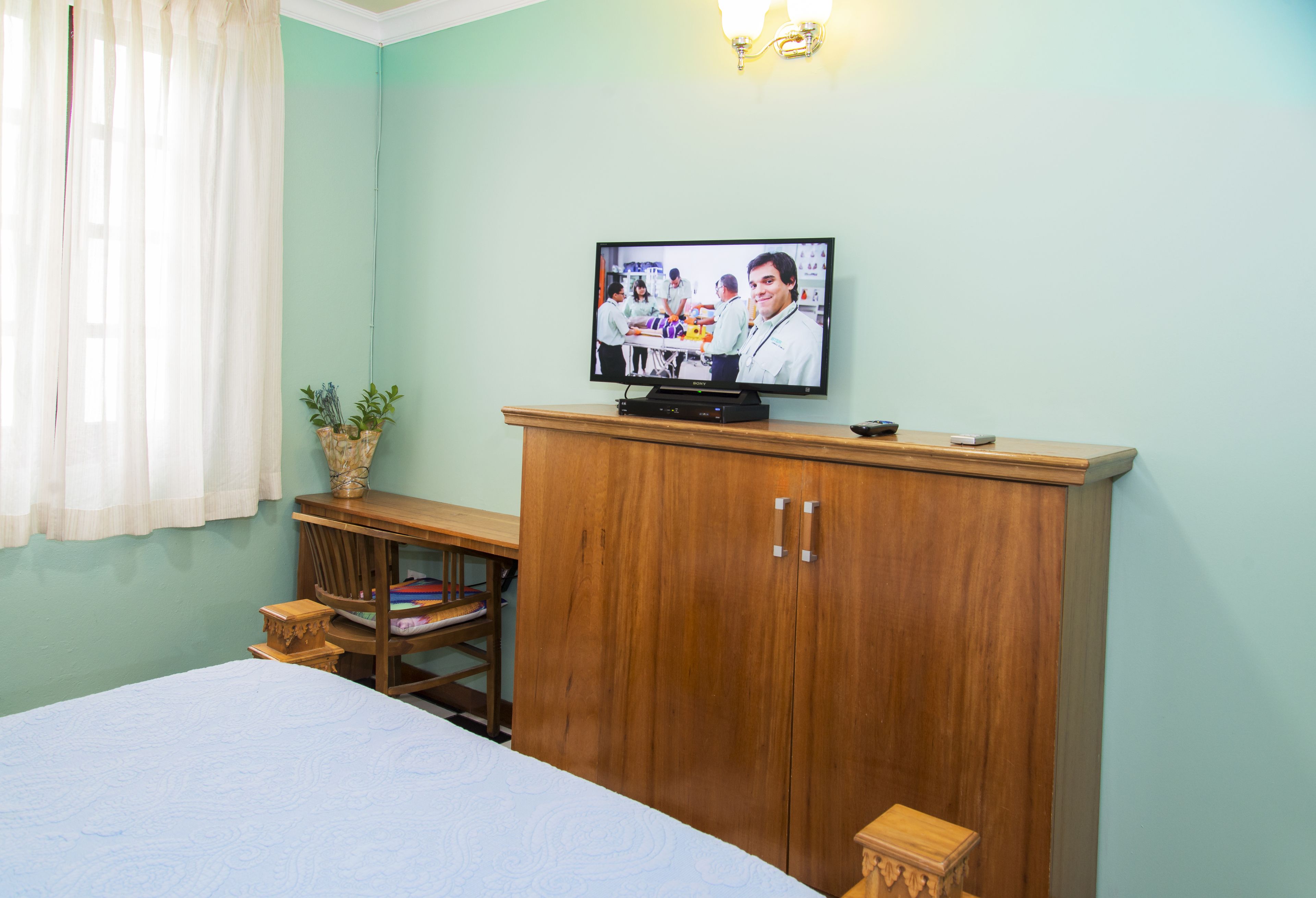 A room with a bed, a wooden wardrobe, a television on top, and a small table with a plant.