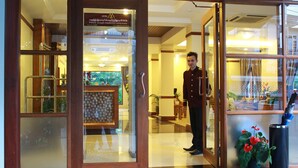 Entrada de um hotel com recepcionista, balcão de atendimento e plantas em vasos.