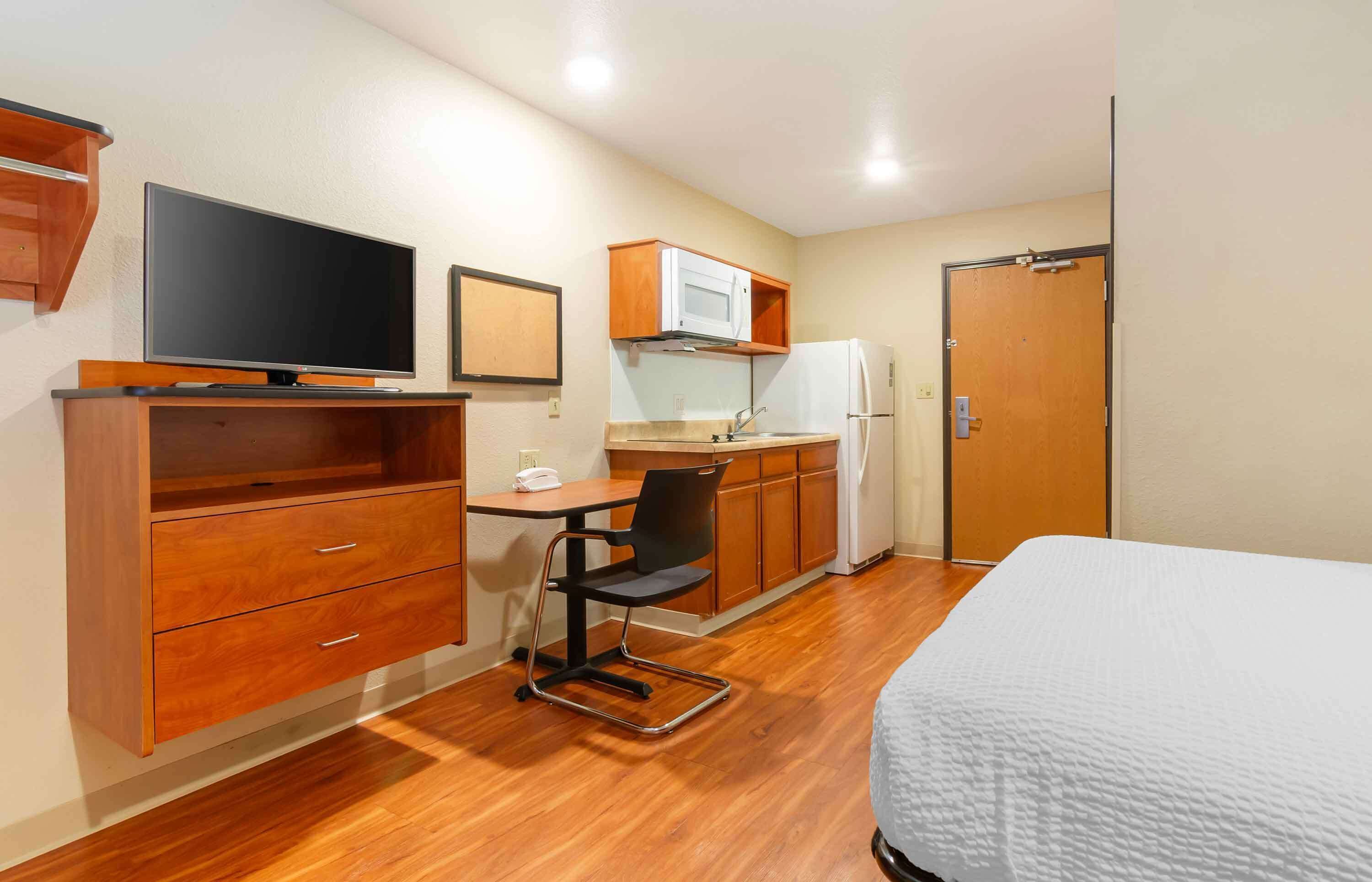 A compact hotel room with a bed, a desk with a chair, a television, a microwave, a refrigerator, and wooden cabinets.
