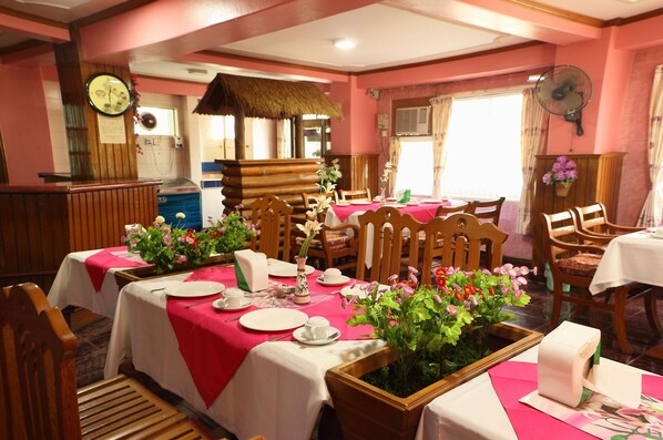 A dining area with tables set for service, wooden chairs, and a thatched roof structure in the background.