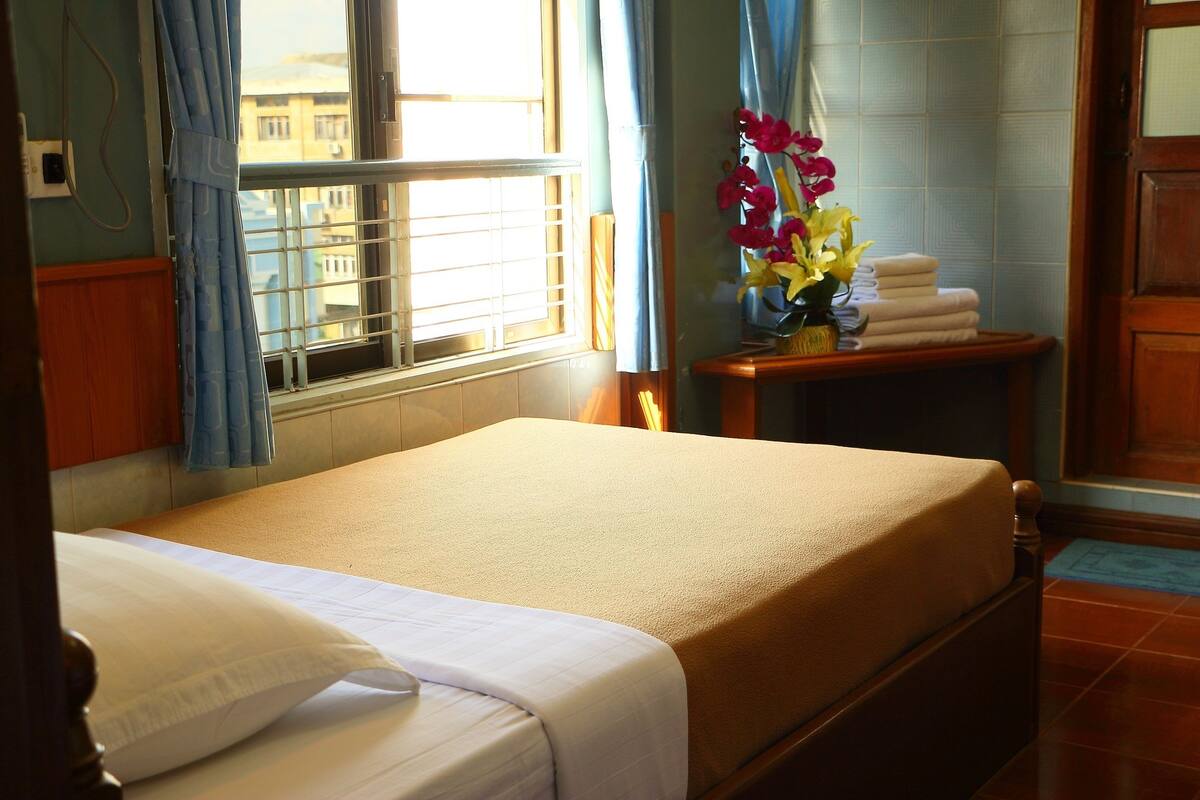A hotel room with a bed, a window with curtains, a shelf with towels and flowers, and a door.