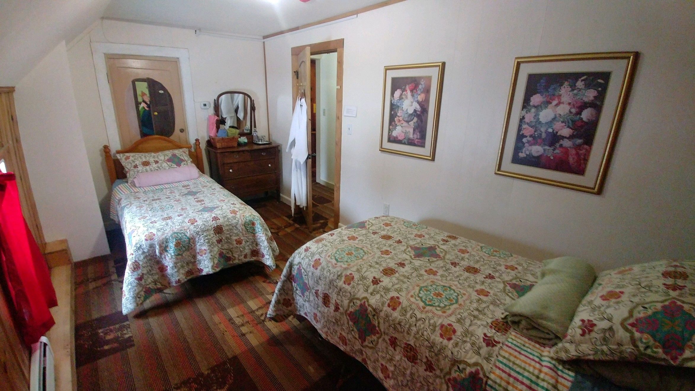 Une chambre avec deux lits, une commode, un miroir et deux tableaux représentant des motifs floraux, encadrés et accrochés au mur.