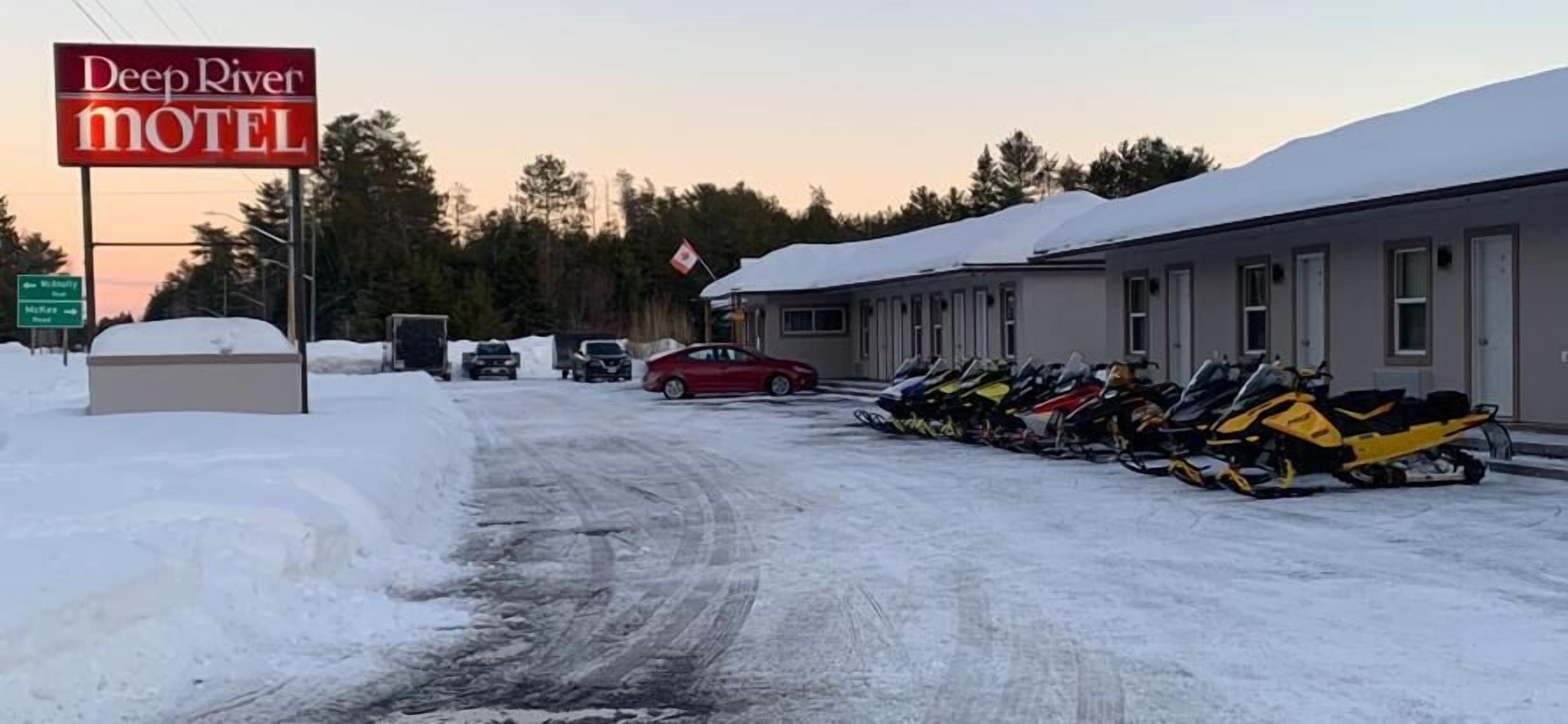 A motel with a sign reading "Deep River Motel," snow-covered ground, and a row of snowmobiles parked outside.