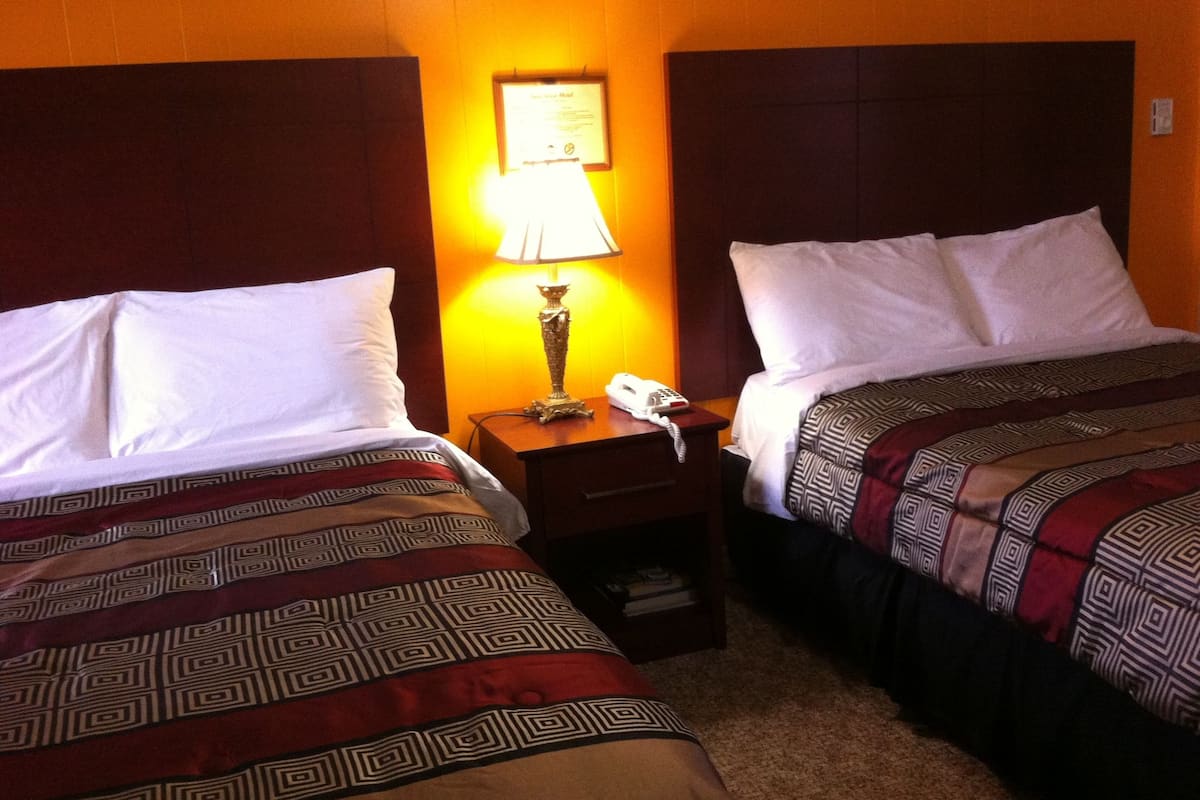 A hotel room with two beds, each with a patterned bedspread, a bedside lamp, a telephone, and a small nightstand with a book.