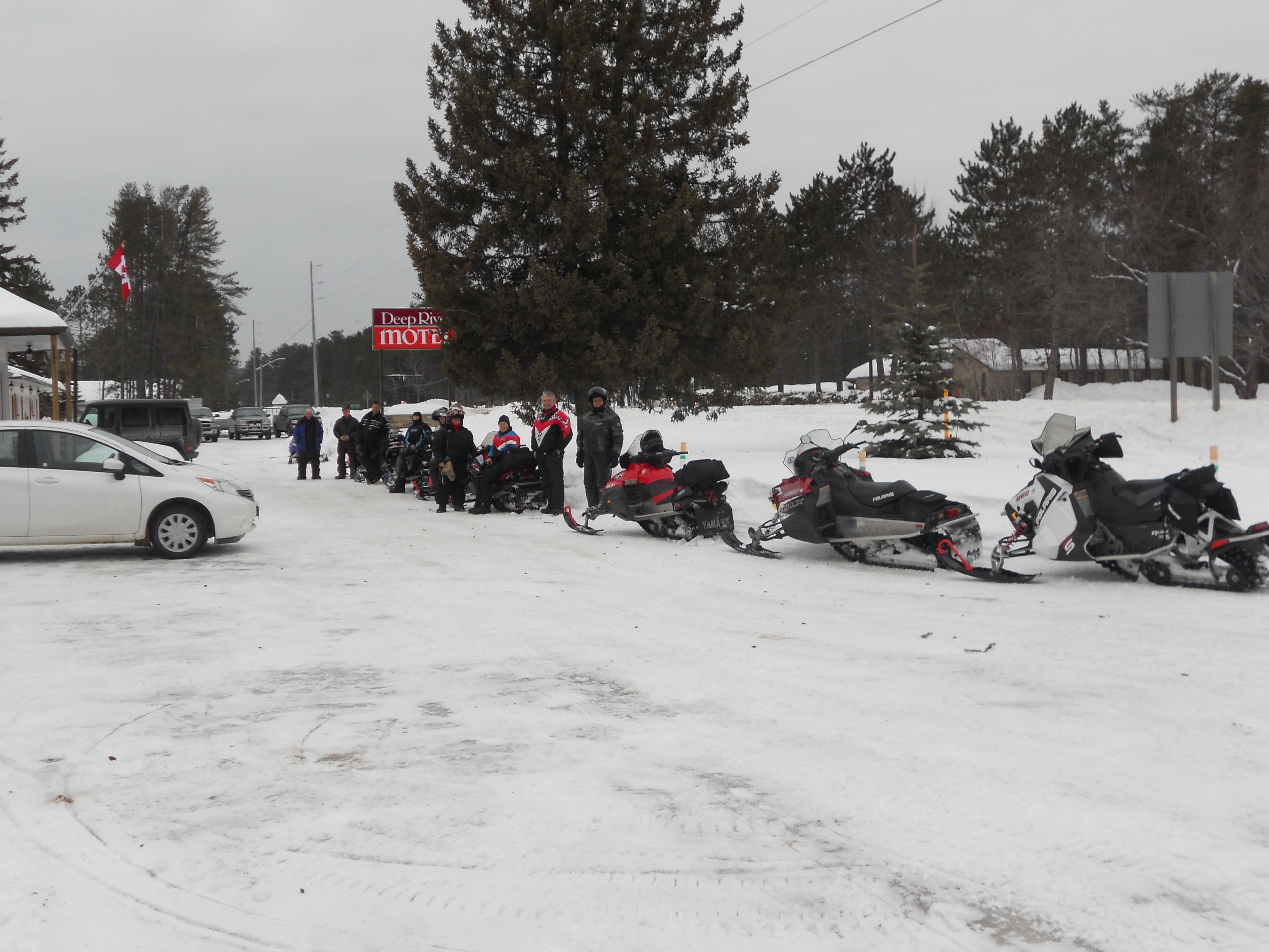 A snow-covered street with parked snowmobiles and a white car.