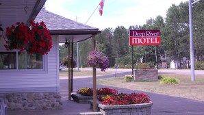 A motel named Deep River with a stone planter with flowers, a hanging flower basket, and a flag.