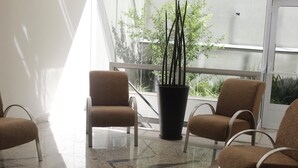 A waiting area with four brown chairs, a large potted plant, and a view of greenery outside through a large window. - Floresta Hotel - Expo Anhembi (São Paulo)