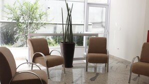 A waiting area with four brown chairs, a large potted plant, and a view of greenery outside through a glass door. - Floresta Hotel - Expo Anhembi (São Paulo)