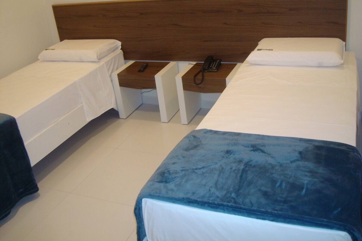 A hotel room with two single beds and a larger bed. There are bedside tables with phones and a power outlet.