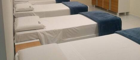 Habitación de hotel con cuatro camas individuales, cada una con colchón blanco y cabecero azul. Se observa un armario de madera adosado a la pared y un equipo de aire acondicionado instalado en el techo.