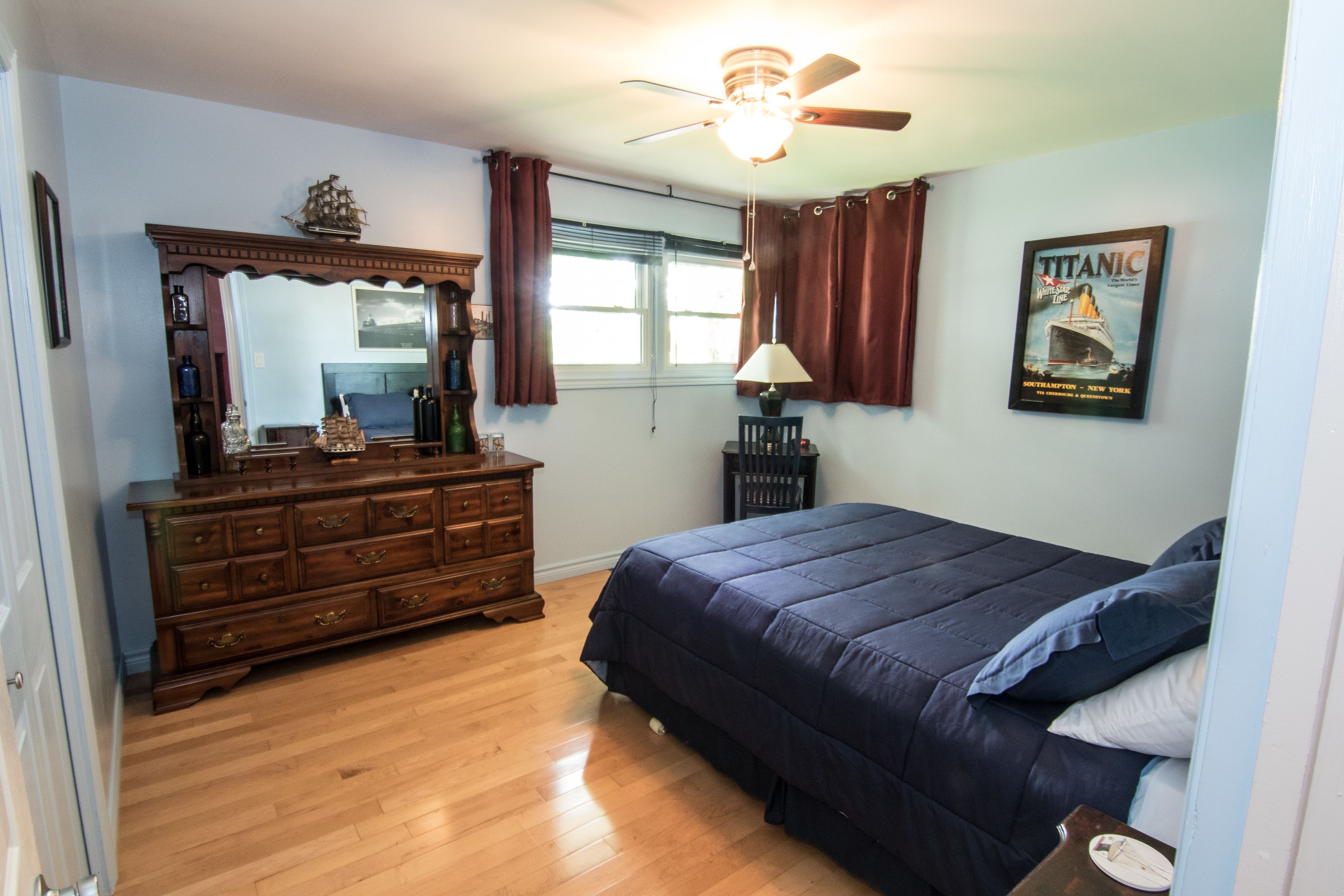 A bedroom with a wooden dresser, a bed with a blue comforter, a ceiling fan, a window with curtains, and a poster on the wall.