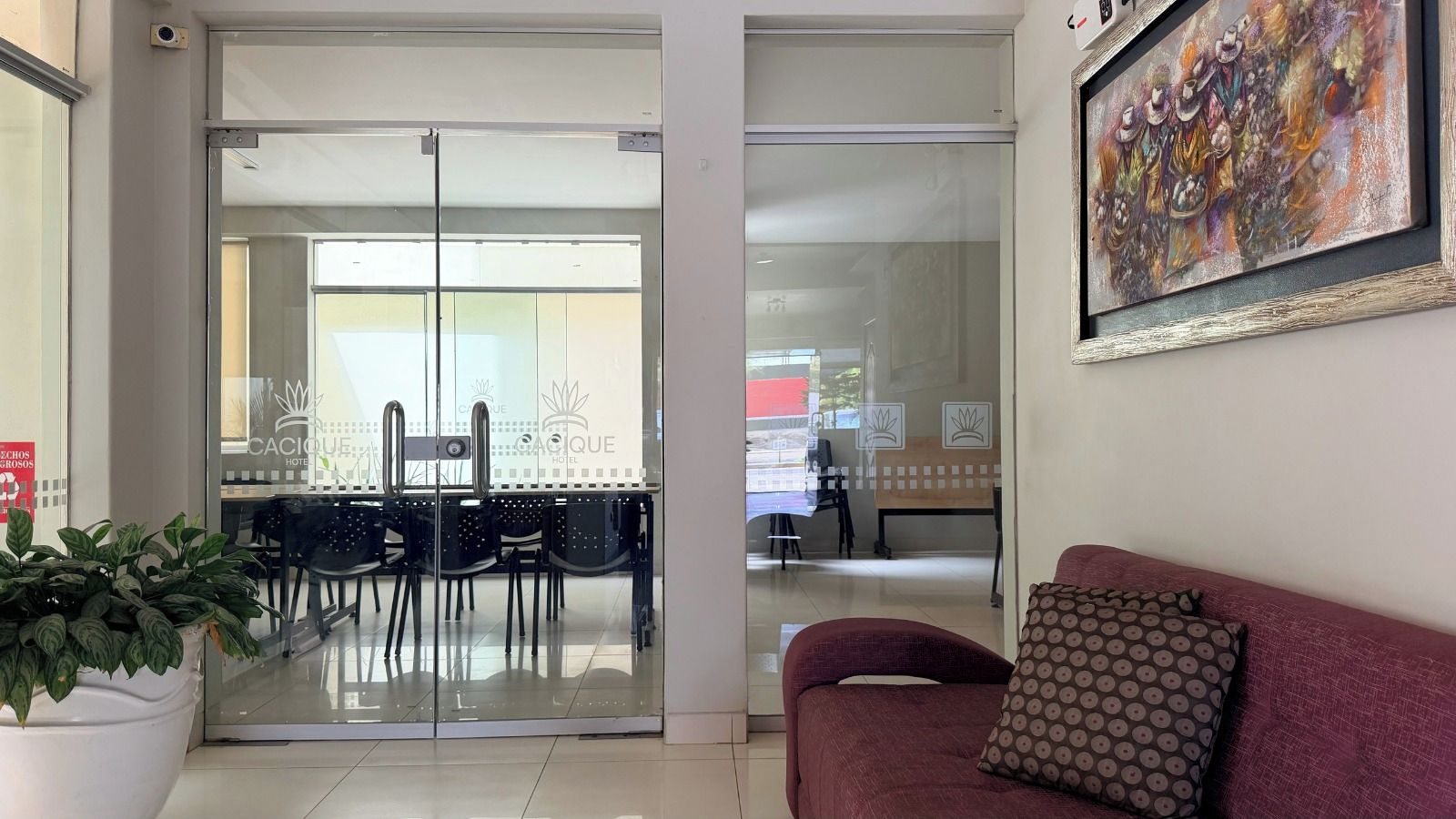 Um hall de hotel com entrada por porta de vidro, área de refeições com cadeiras e mesas, sofá com almofadas estampadas e um quadro abstrato emoldurado na parede.