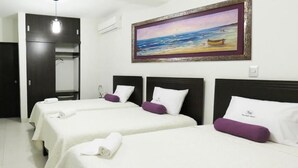 A hotel room with two beds, each with white linens and purple pillows, a painting of a beach scene on the wall, and a door leading to a bathroom. - El Cacique Hotel (Piura)