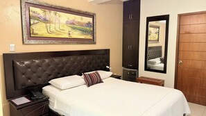 A bedroom with a large bed, a nightstand, a wardrobe, a mirror, and a painting on the wall. - El Cacique Hotel (Piura)