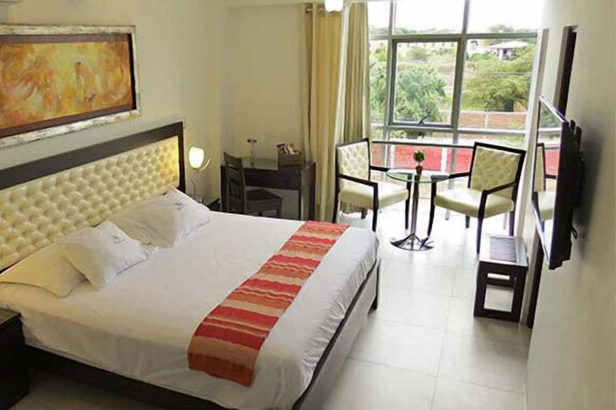 A hotel room with a large bed, a bedside table with a lamp, a painting on the wall, and a balcony with chairs and a table.