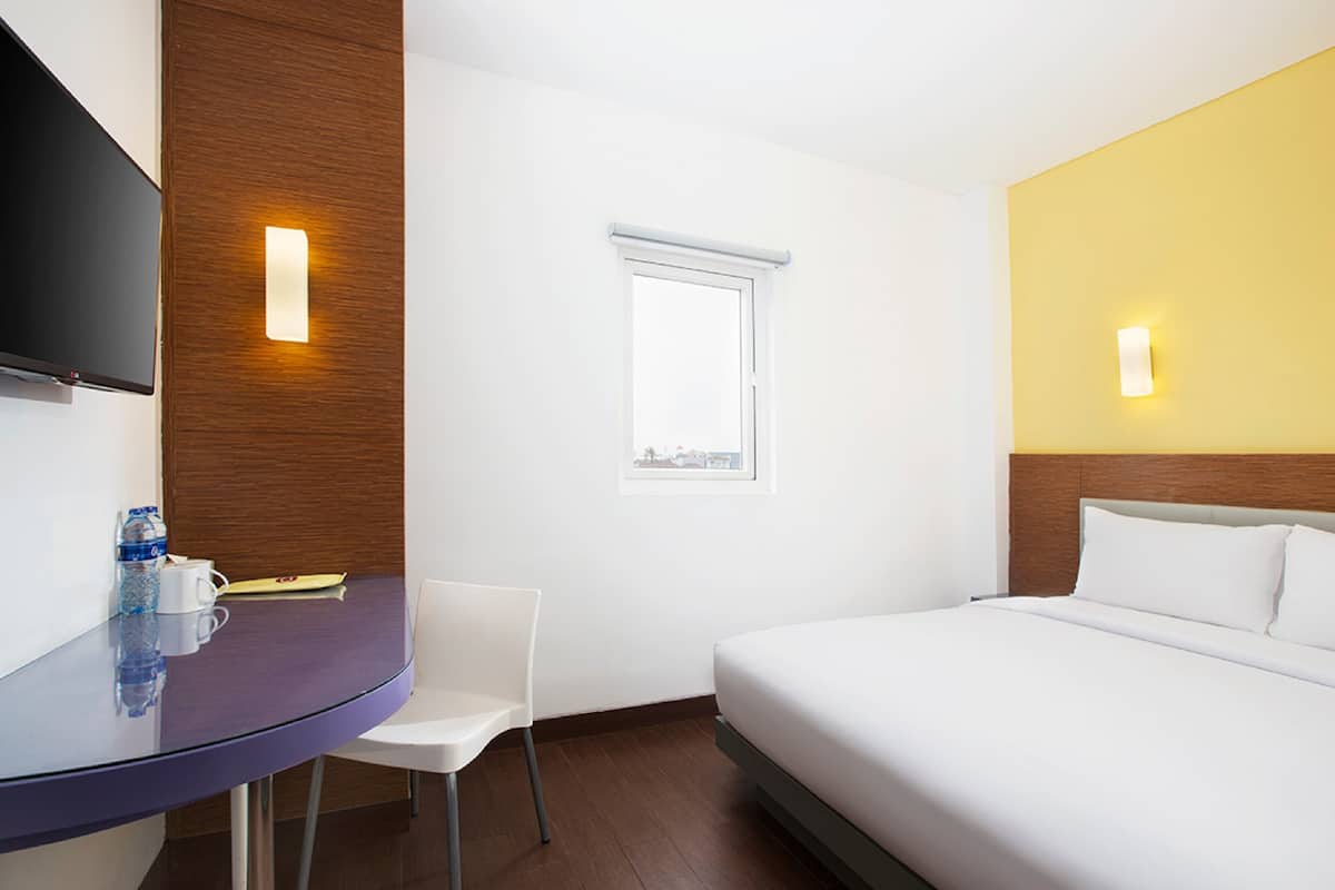 A hotel room with a bed, a desk with a chair, a television mounted on the wall, and a window.