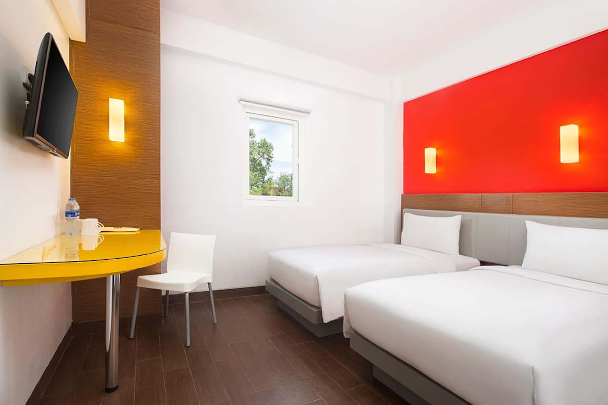 A hotel room with two beds, a small table with a chair, a TV mounted on the wall, and a window with a view of trees.