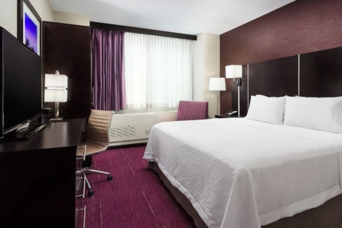 Quarto de hotel com uma cama grande, uma mesa de escritório com um computador, uma cadeira, uma televisão e uma janela com cortinas.