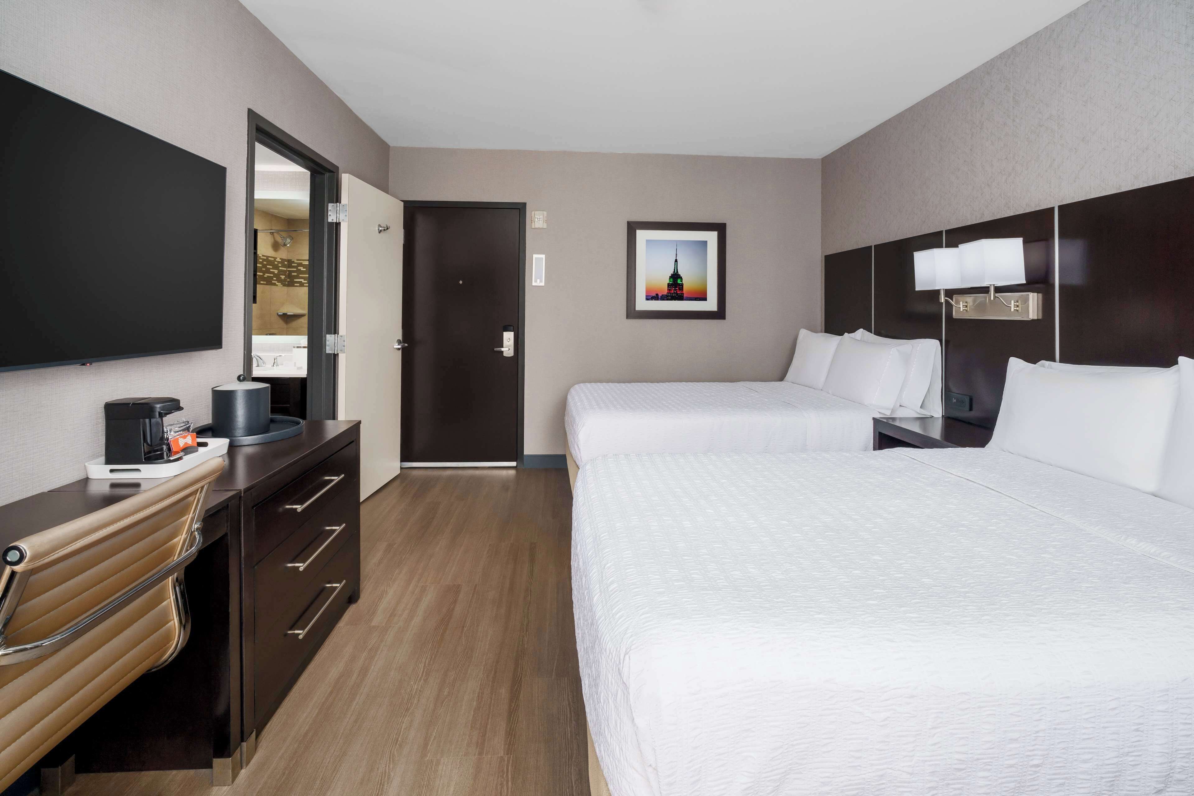Quarto de hotel com uma cama grande, uma cama menor, uma mesa com cafeteira, uma cadeira, uma televisão e um quadro pendurado na parede.