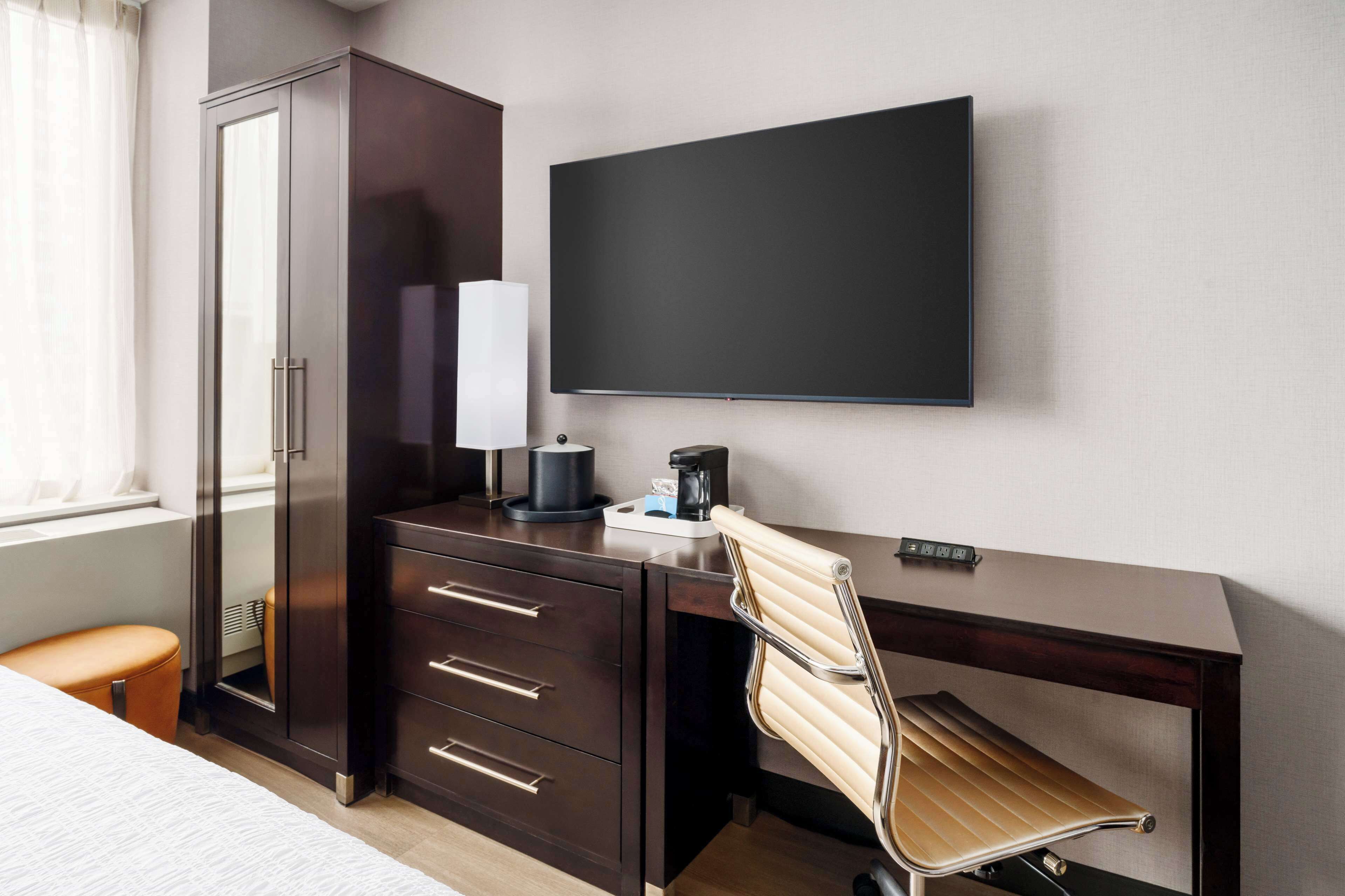 Habitación de hotel con cama, escritorio con silla, armario, televisor de pantalla plana y cafetera.