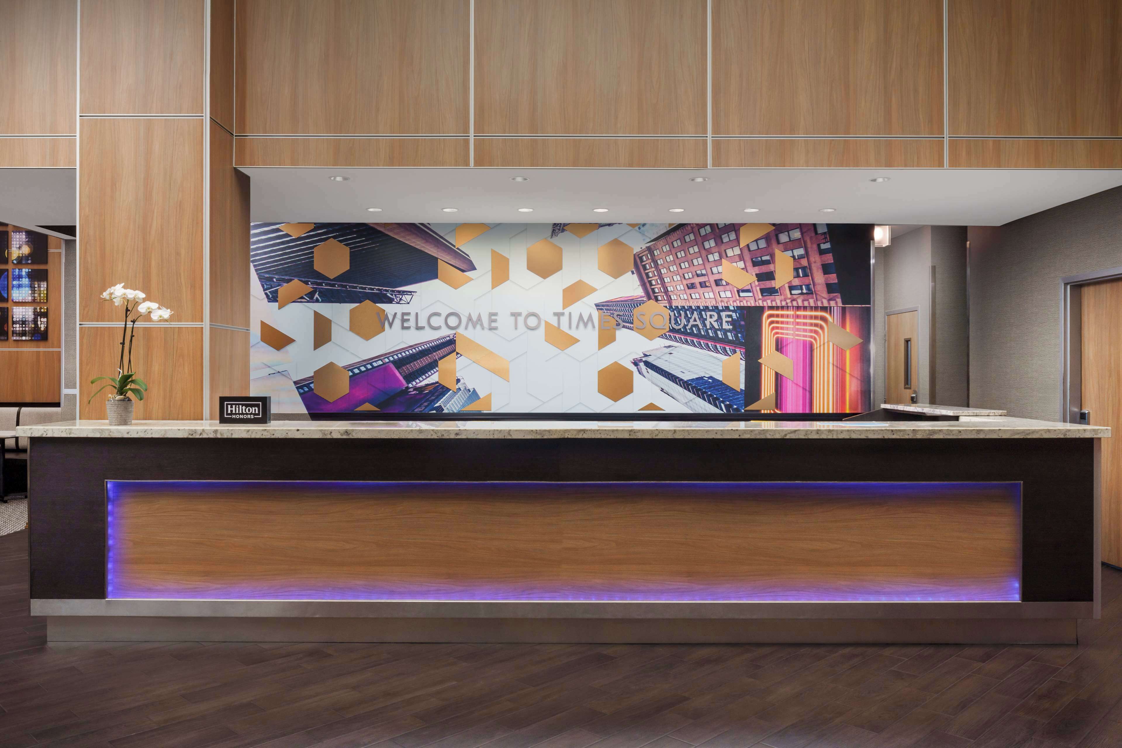 A modern hotel reception desk with a "Welcome to Times Square" mural, a "Hilton" sign, and a purple-lit reception area.