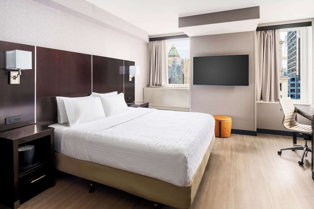 Une chambre d’hôtel dotée d’un grand lit, d’un téléviseur à écran plat, d’un bureau avec une chaise et offrant une vue sur le paysage urbain par la fenêtre.