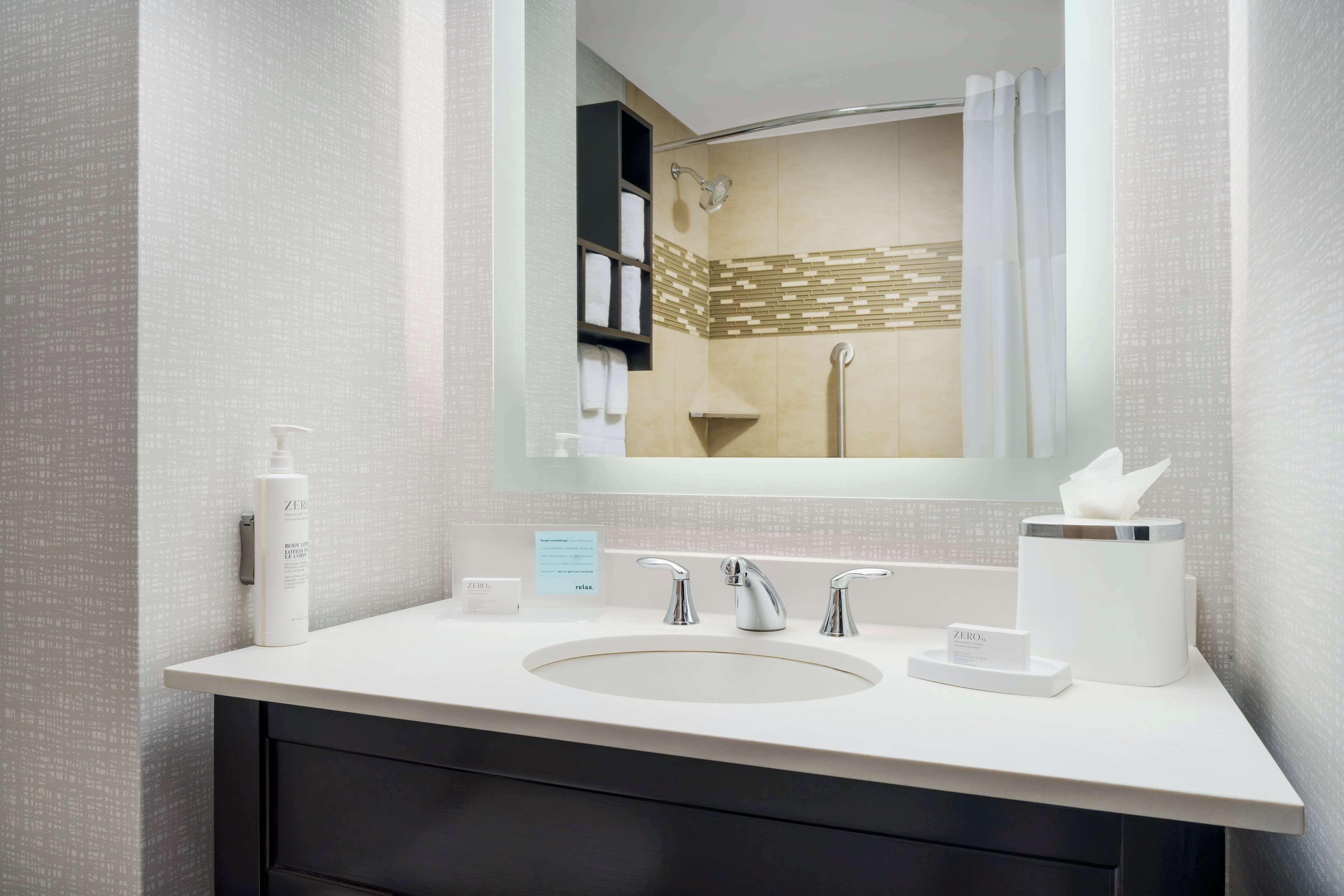 A modern bathroom with a large mirror, a sink with two faucets, a towel dispenser, and a shower area visible in the mirror reflection.