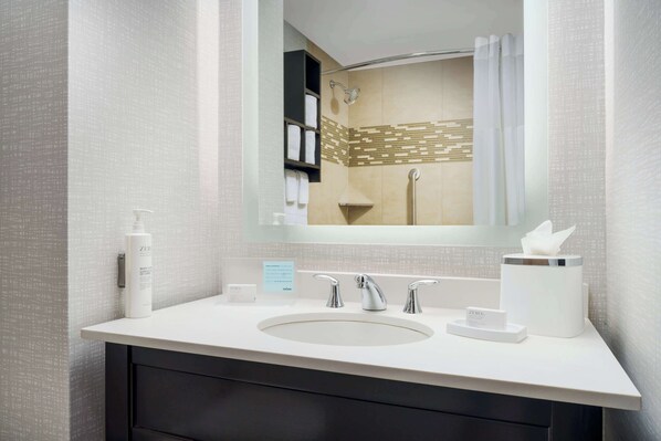 A modern bathroom with a large mirror, a sink with two faucets, a towel dispenser, and a shower area visible in the mirror reflection.