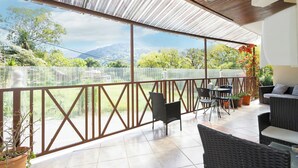 Un patio techado con mobiliario exterior, vistas a la vegetación y montañas, y una barandilla metálica.