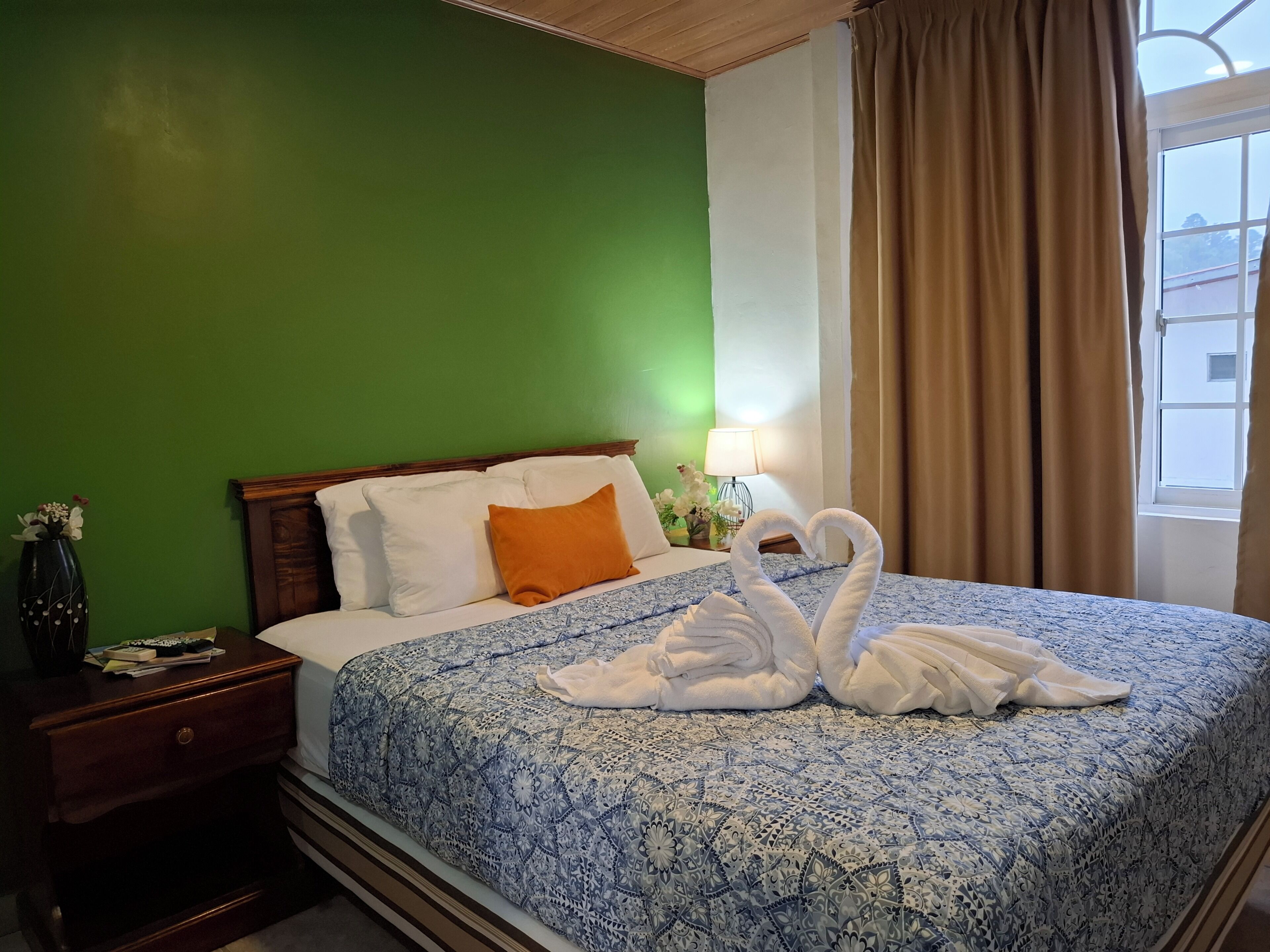 A hotel room with a bed, green walls, a wooden headboard, a nightstand, a lamp, and a window with curtains.