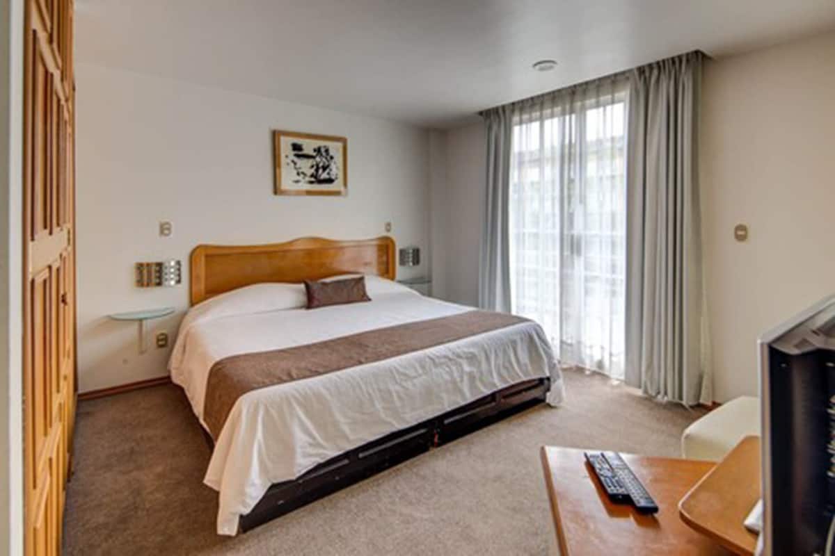 Une chambre d’hôtel avec un lit simple, un bureau avec un ordinateur, une télévision, une petite table de chevet et une fenêtre avec des rideaux.