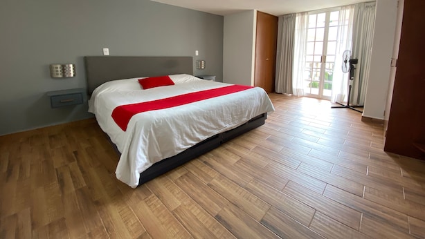 A modern hotel room with a large bed, wooden flooring, a grey wall, and a large window with curtains.