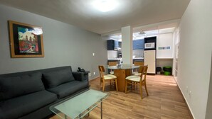 A living room with a dark sofa, a glass coffee table, and a dining area with a round table and chairs. A kitchen is visible in the background with cabinets and appliances.