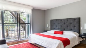 A modern hotel room with a large bed, a grey upholstered headboard, a red accent pillow, and a view of a tree through a sliding glass door.