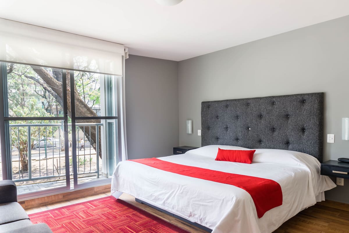 A modern hotel room with a large bed, a grey upholstered headboard, a red accent pillow, and a view of a tree through a sliding glass door.