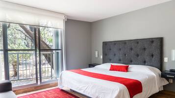Habitación de hotel moderna con una cama grande, cabecera tapizada en gris, almohada decorativa roja y vista a un árbol a través de una puerta corrediza de vidrio.