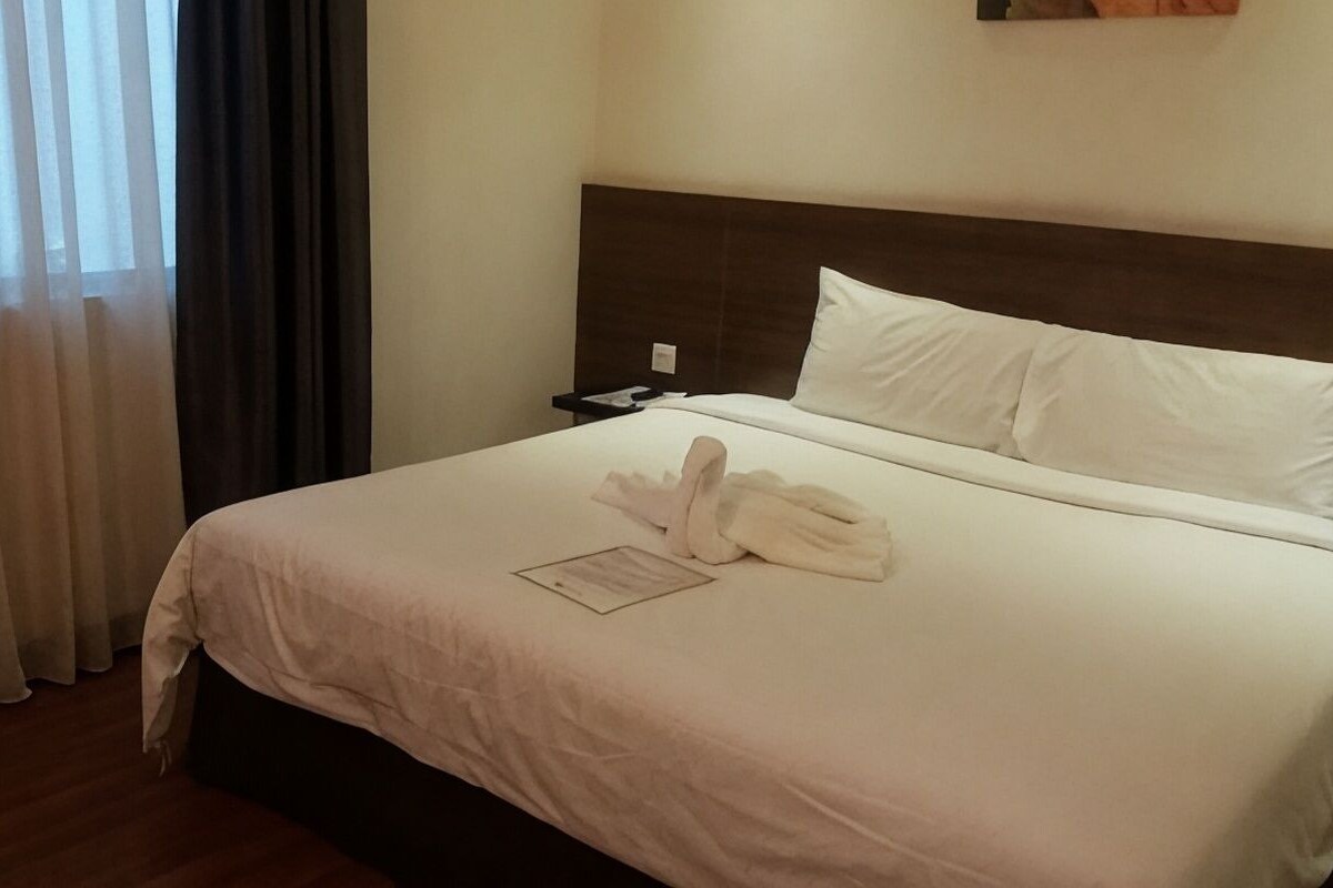 A hotel room with a neatly made bed, a nightstand, a wall-mounted phone, and a window with curtains.