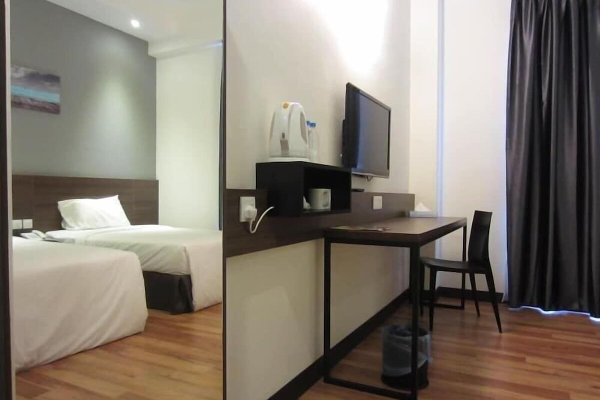 Quarto de hotel com uma cama, escrivaninha com cadeira, televisão e janela com cortinas.