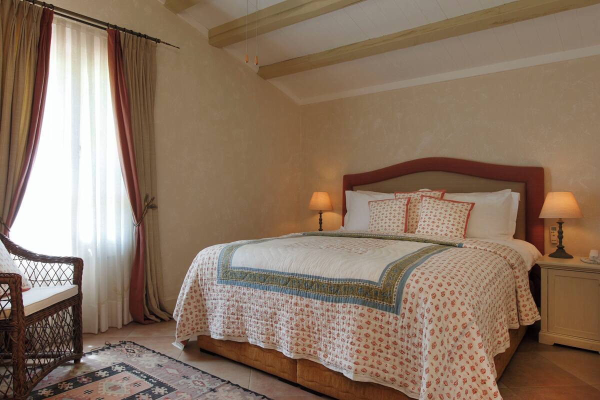 Un dormitorio con una cama grande, dos lámparas de noche, un cabecero de madera, una silla de mimbre y una alfombra estampada.