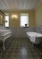 Un baño con una bañera independiente, un tocador con espejo y un espejo redondo, una ventana con cortinas y un suelo estampado.