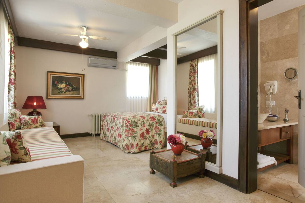 Habitación de hotel con cama, sofá, una mesita con flores, espejo y baño con lavabo y toallero.