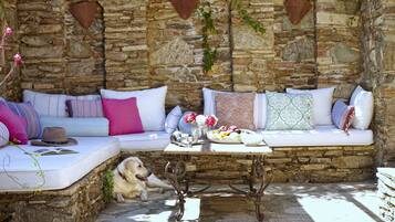 Un espace extérieur aménagé avec des murs en pierre, doté de bancs rembourrés et d’une table ornée de fleurs et de fruits.