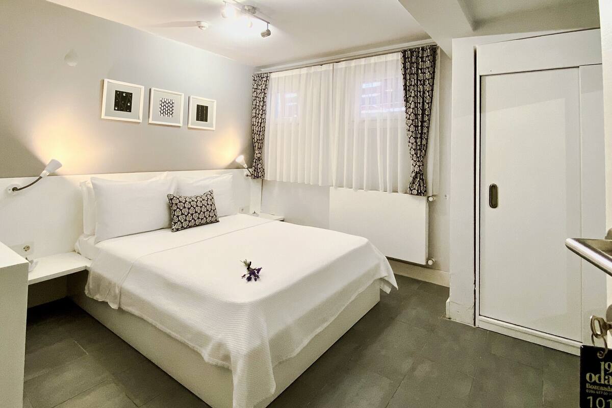 A bedroom with a bed, a nightstand, a wall-mounted lamp, a window with curtains, and a door.