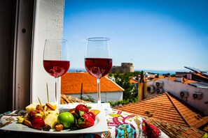 A table with a plate of fruit and nuts, two glasses of red wine, and a view of a cityscape with buildings and a clear sky. - 19 Oda Bozcaada (Adults Only) (Bozcaada)